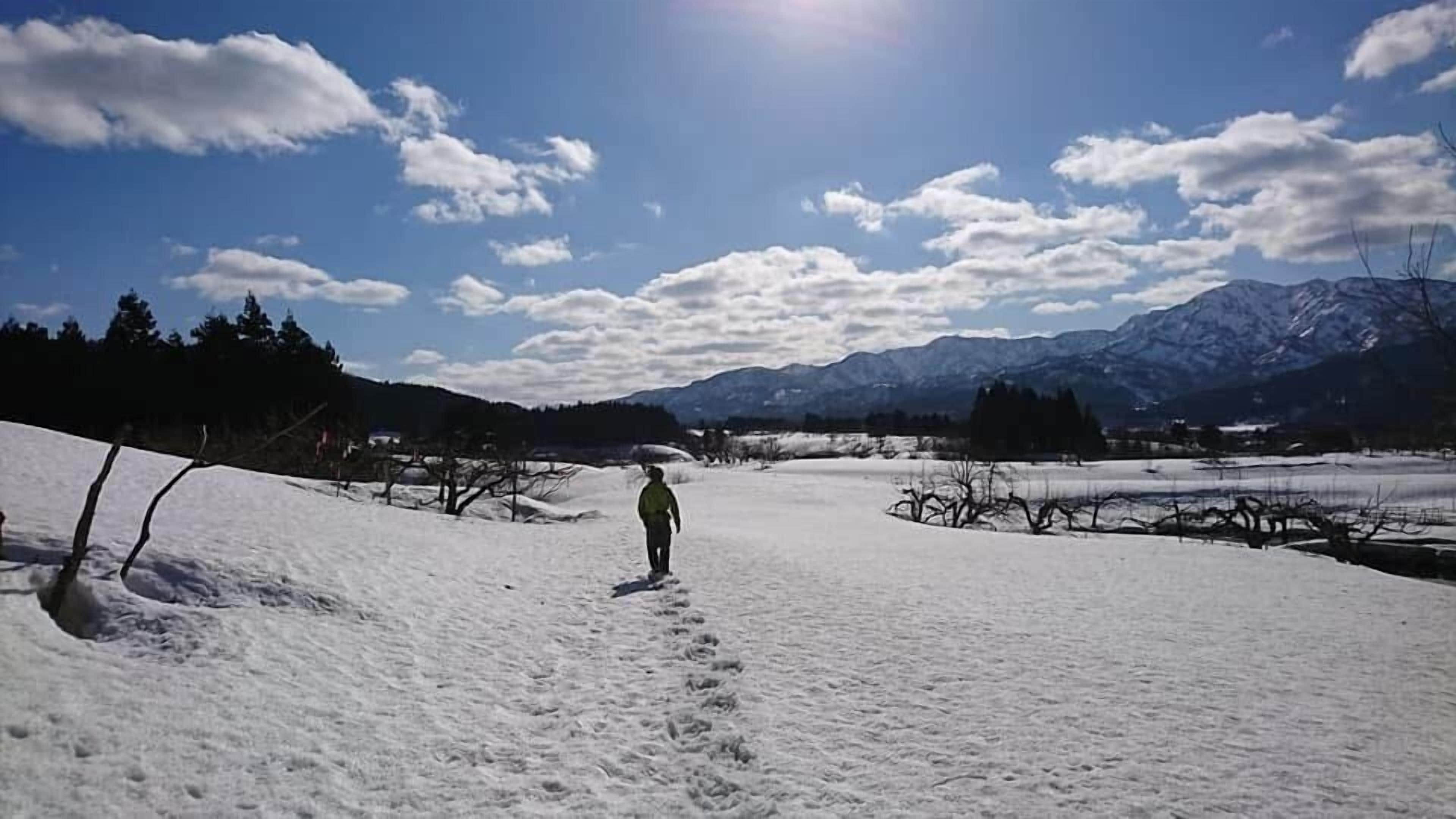 A snowy landscape with a person walking on a path, mountains in the background, and a clear sky.
