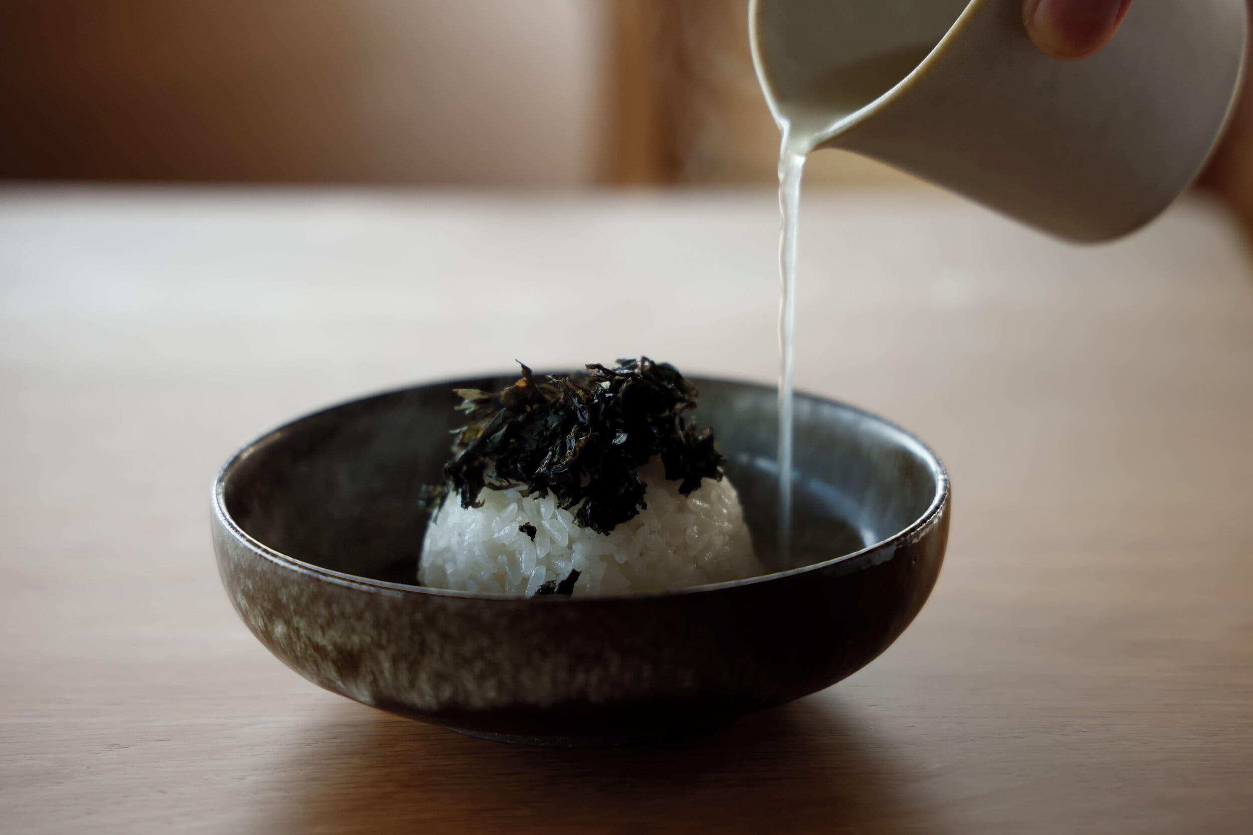 A bowl of white rice topped with seaweed, with a liquid being poured over it.