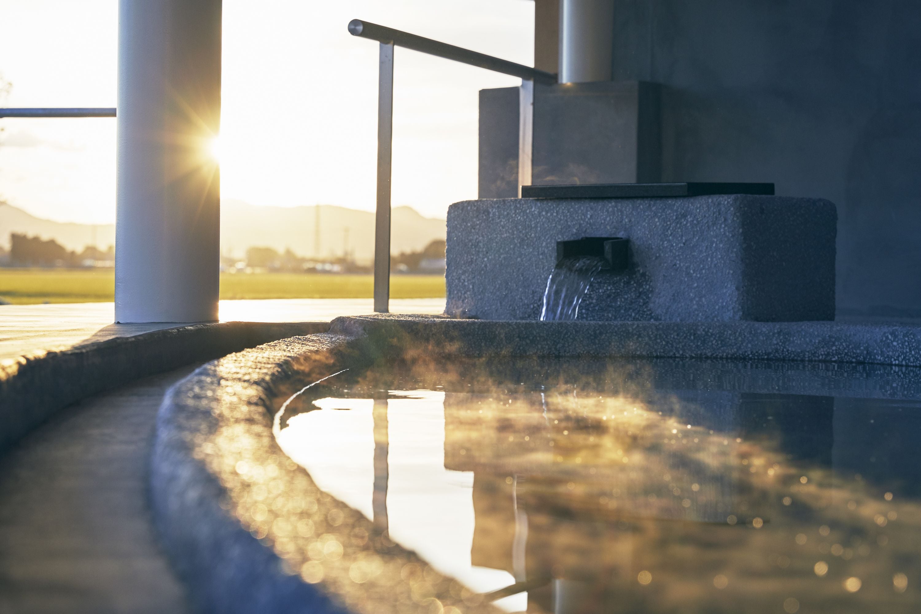 A water feature with a metal railing and a stone structure, set against a backdrop of a sunset.
