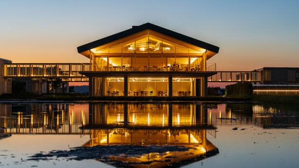 A modern building with a glass facade reflecting in a body of water, featuring a flat roof and multiple levels.