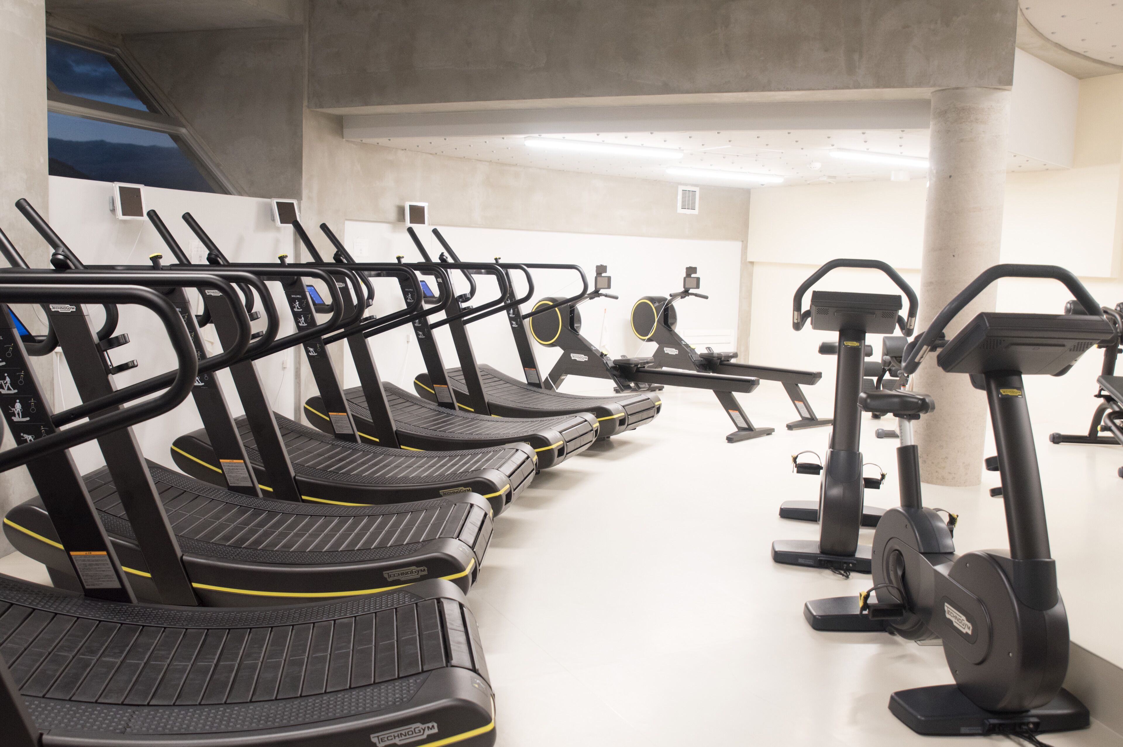 Filas de bicicletas estáticas y cintas de correr en un gimnasio con paredes blancas y suelo de hormigón.