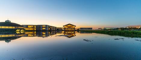 A modern building with a glass facade reflecting in a calm body of water, surrounded by greenery.