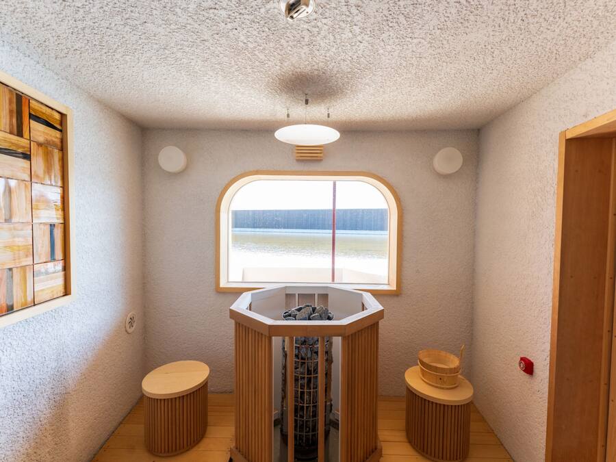 A small, cozy room with a large window, a wooden stool, a small table, and a wooden cabinet.