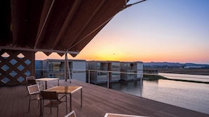 A wooden deck with tables and chairs, overlooking a body of water and a sunset.