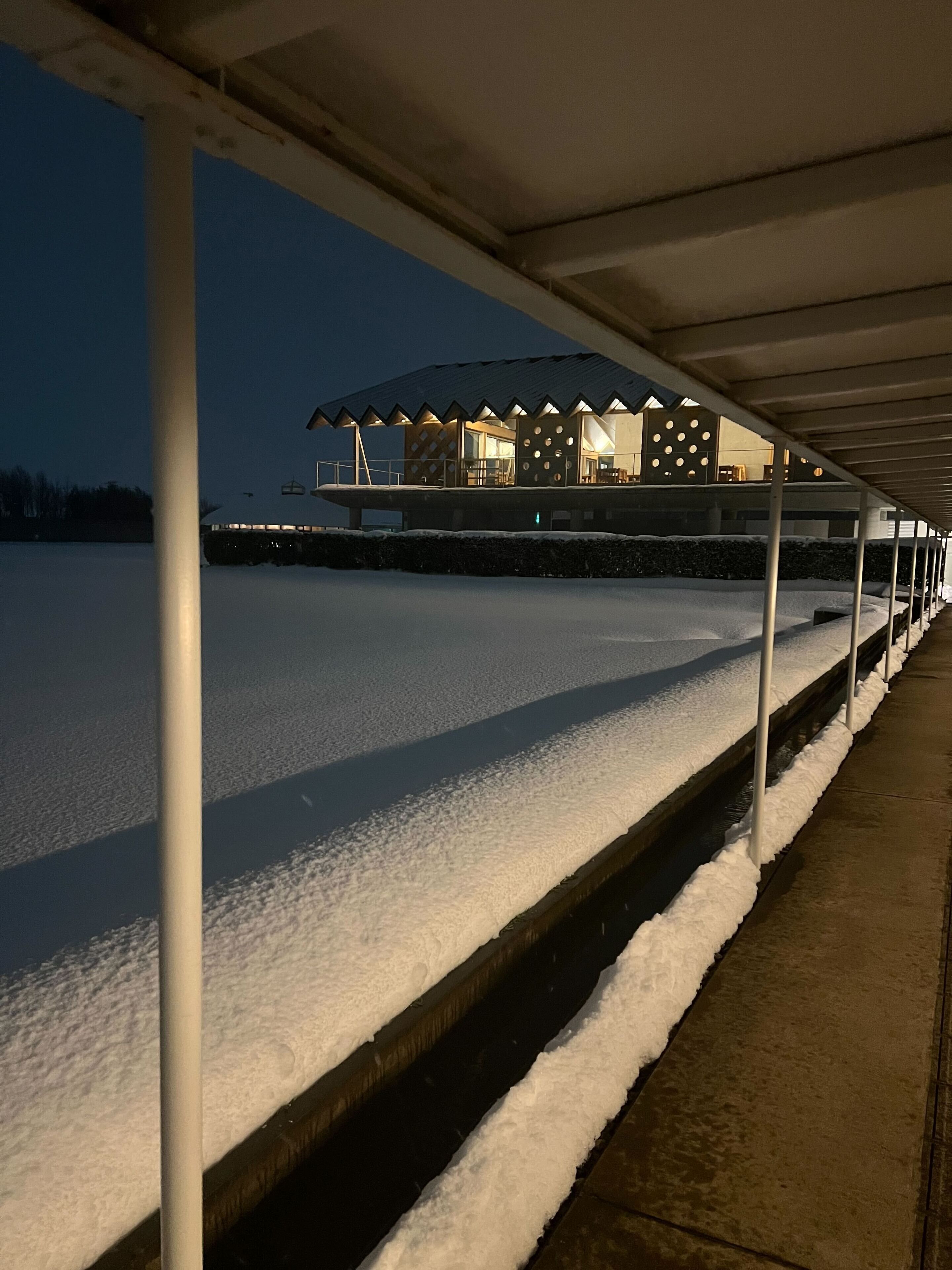 A covered outdoor area with a snow-covered ground, a building with a modern design featuring circular windows, and a metal structure in the foreground.