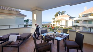 A balcony with a table set for two, a chair, a sun lounger, and a view of the sea and buildings. - 213 Lux-Serv Apart-Estepona, 2-B'room, All Year 28c, 20m Heated Pool. Sea Views (Estepona)