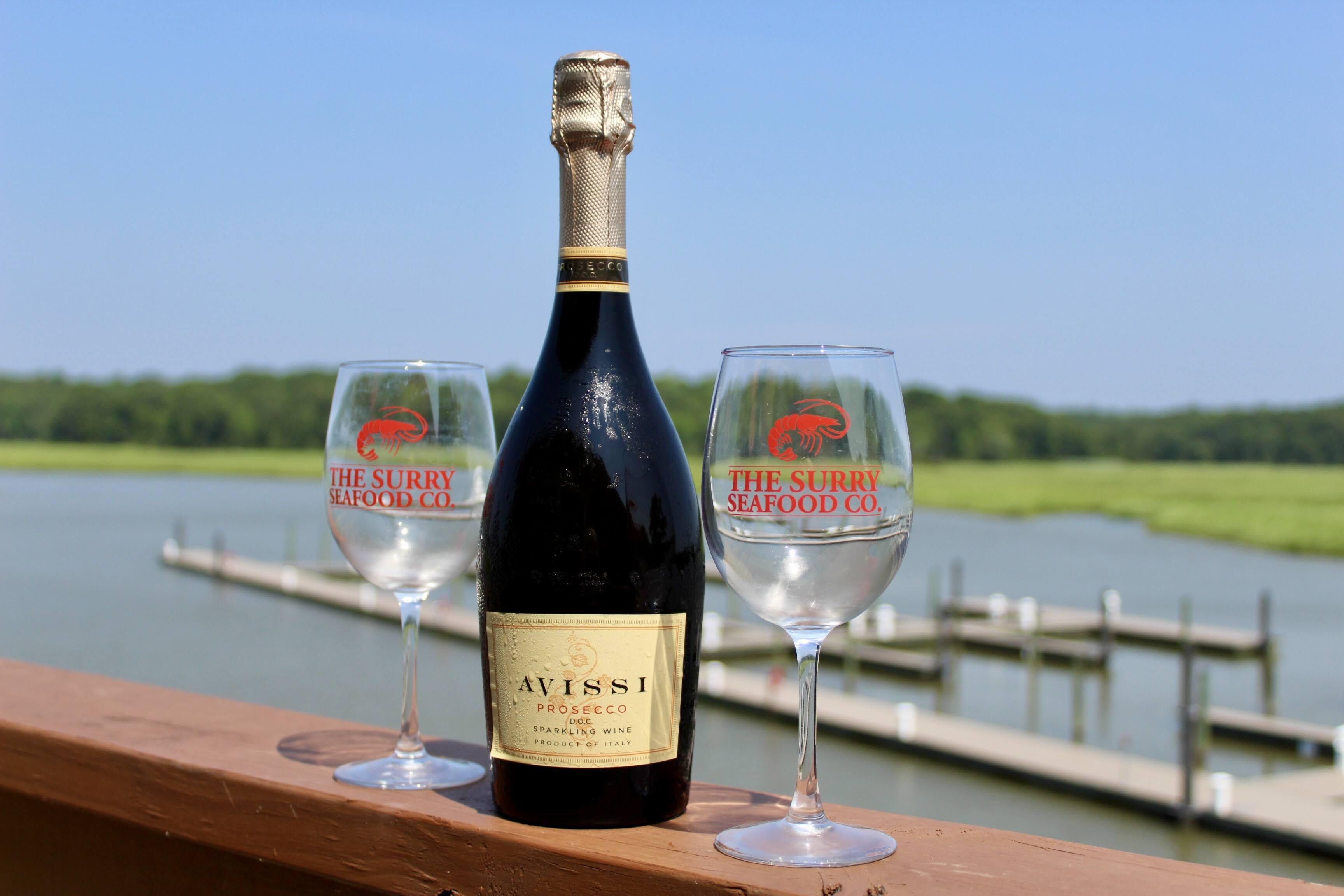 A bottle of Avissi Prosecco DOC sparkling wine and two glasses with the Surry Seafood Co. logo on a wooden deck overlooking a body of water.