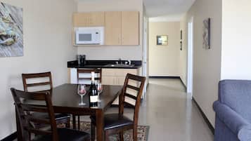 Un comedor con una mesa de madera, cuatro sillas y una botella de vino. Se ve una cocina con microondas y armarios al fondo.