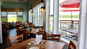 A restaurant with wooden tables and chairs, large windows, and a view of a green field.