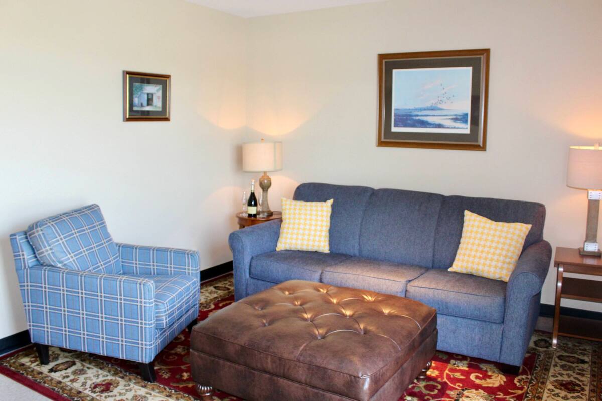 A living room with a blue armchair, a blue sofa with yellow pillows, and a brown ottoman. There are two framed pictures on the walls and a lamp on a side table.