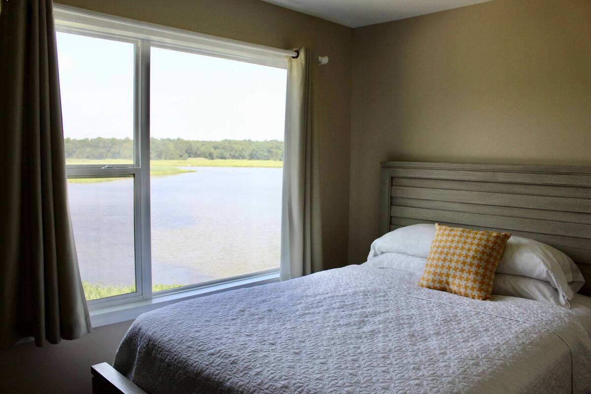 A bedroom with a large window, a bed with white bedding, and a view of a lake.