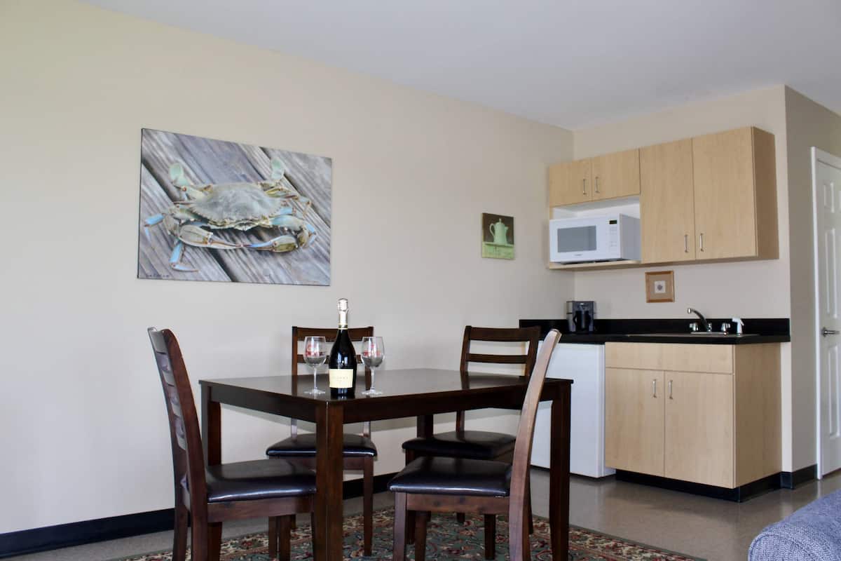 A dining area with a wooden table and chairs, a bottle of wine, and two glasses. There is a microwave and a sink in the adjacent kitchen area.