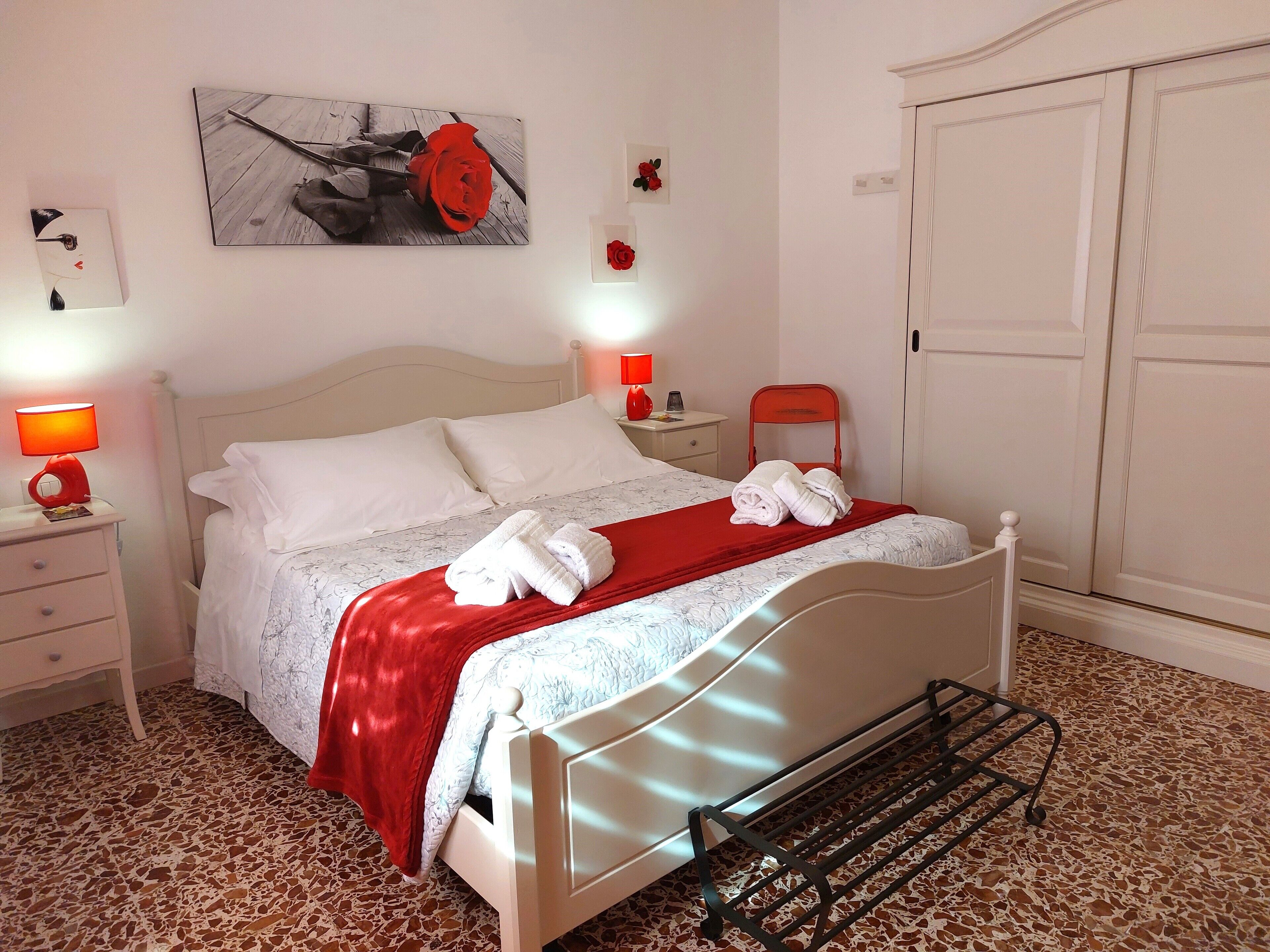 Un lit bien fait, avec du linge de lit blanc et un plaid rouge, une table de chevet avec une lampe, et un tableau représentant une rose accroché au mur.