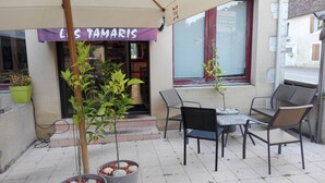 Ulkona on oleskelualue, jossa on pöytä ja tuoleja, pieni puu ja kyltti, jossa lukee »LES TAMARIS».