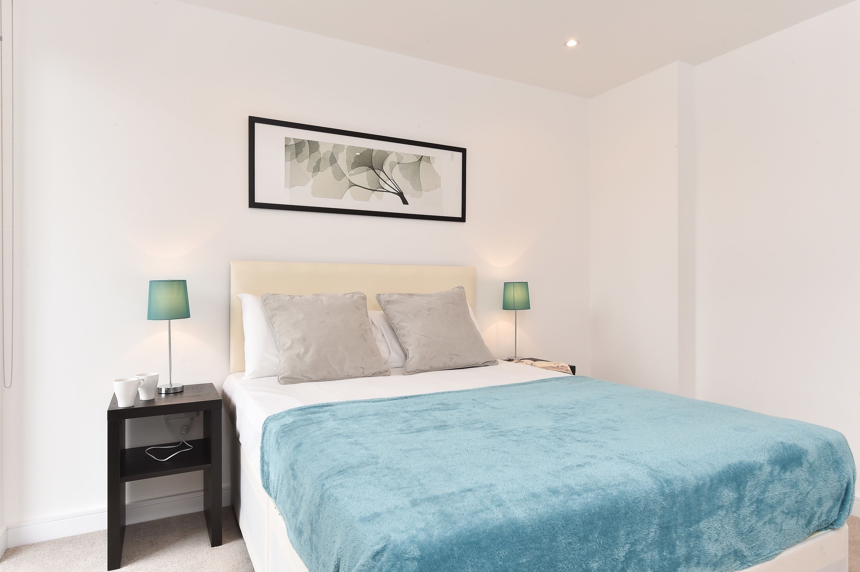 Une chambre moderne avec un lit, deux tables de chevet, une commode, une couverture bleue et une œuvre d’art encadrée accrochée au mur.