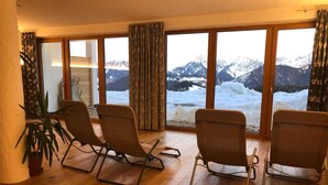 Quarto com quatro poltronas reclináveis, um vaso de planta e uma janela ampla com vista para montanhas cobertas de neve.