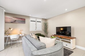 Uma sala de estar moderna com um sofá cinza, uma escrivaninha com luminária, uma televisão sobre um suporte de madeira e uma janela grande com persianas.