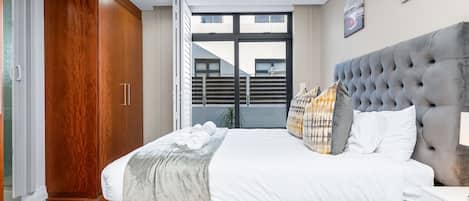 A bedroom with a large bed, a grey upholstered headboard, and a view through a sliding door to an outdoor area.