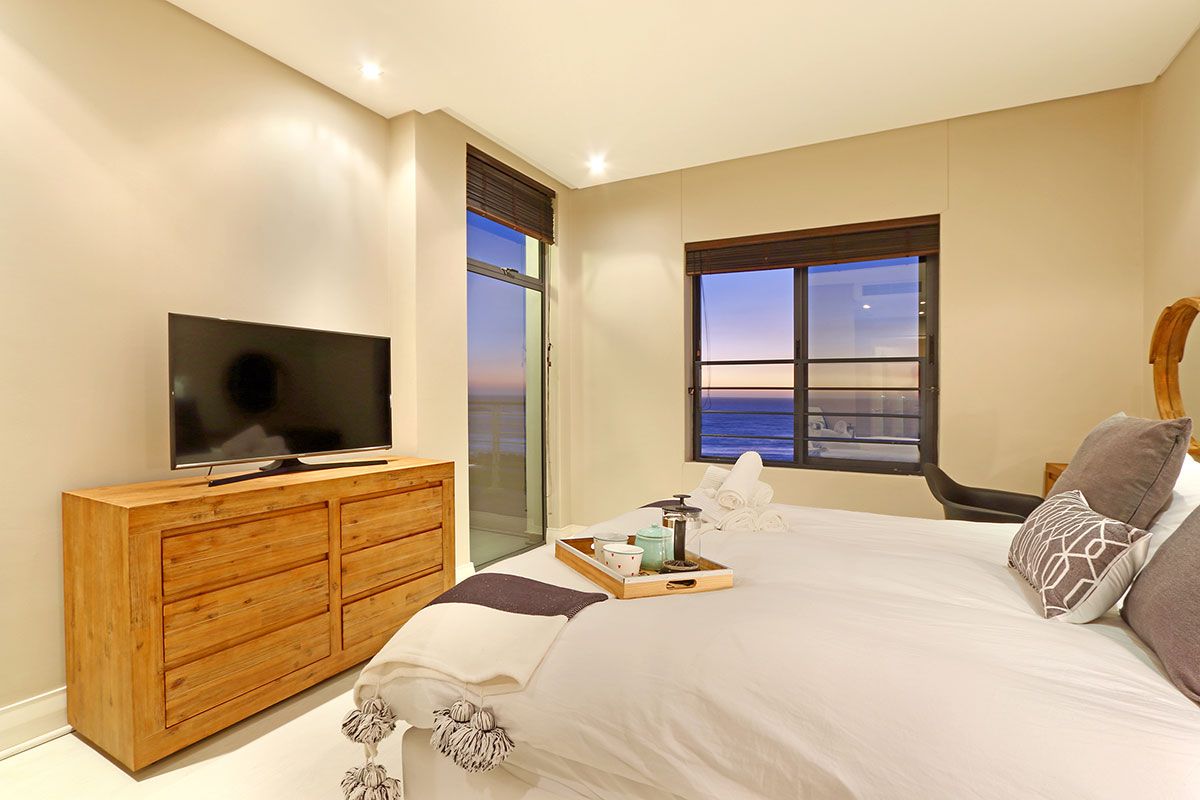 A bedroom with a TV on a wooden dresser, a bed with white bedding, and a window with a view.