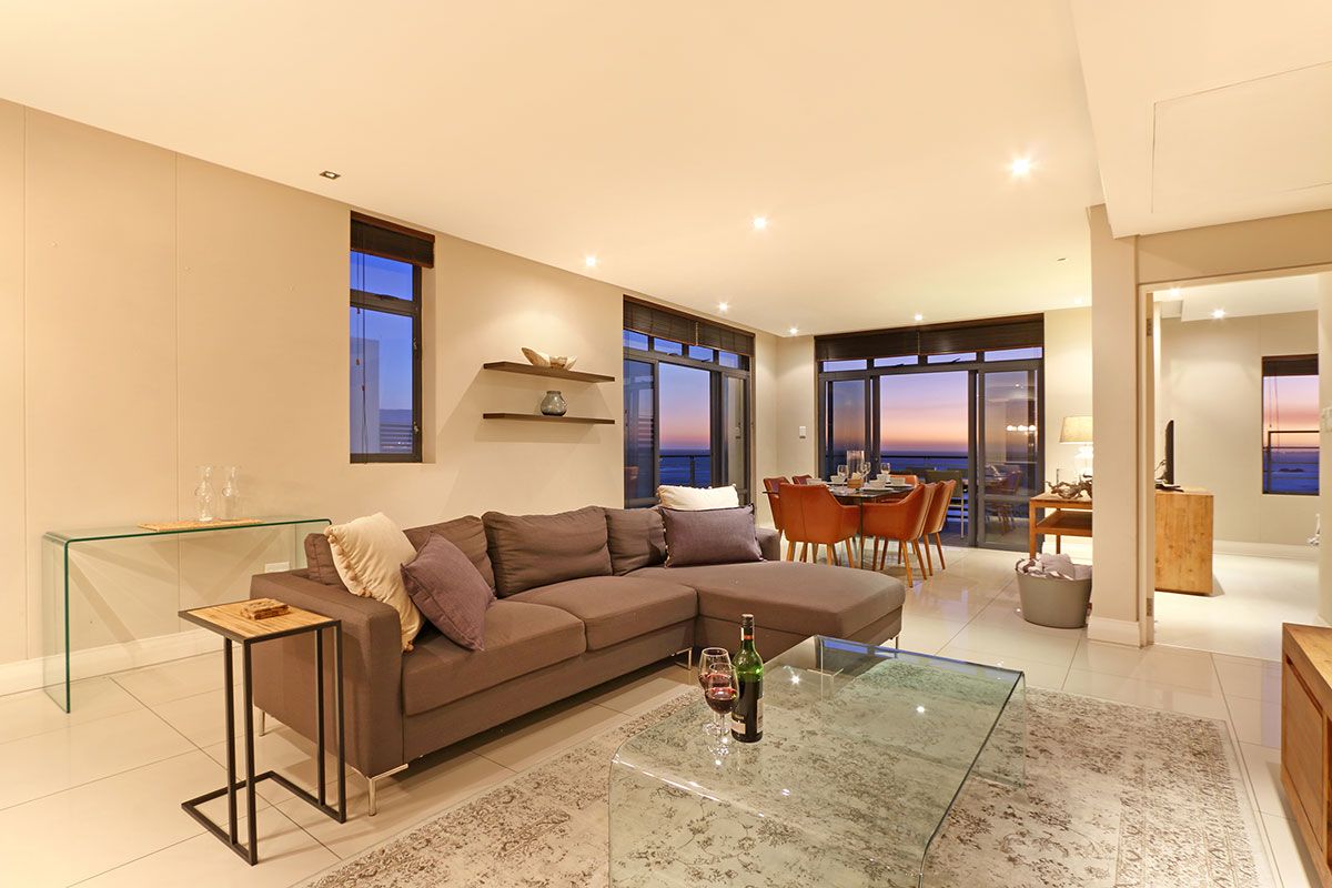 Una sala de estar moderna con una mesa de centro de vidrio, un sofá seccional marrón y un comedor con vistas al atardecer.