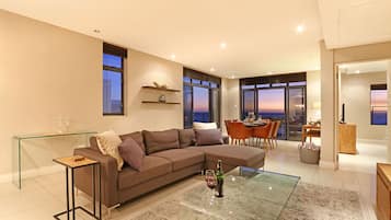 Una sala de estar moderna con una mesa de centro de vidrio, un sofá seccional marrón y un comedor con vistas al atardecer.
