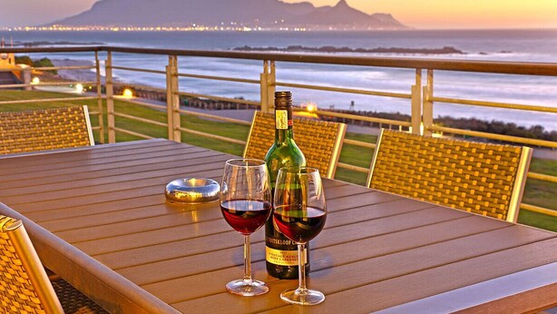 A table with two wine glasses and a bottle of wine, set against a scenic ocean view with a mountain in the background. - Eden on the Bay 277 (Cape Town)