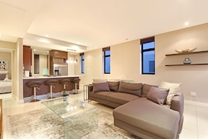 A modern living room with a glass coffee table, a brown sofa, and a kitchen area in the background. - Eden on the Bay 277 (Cape Town)