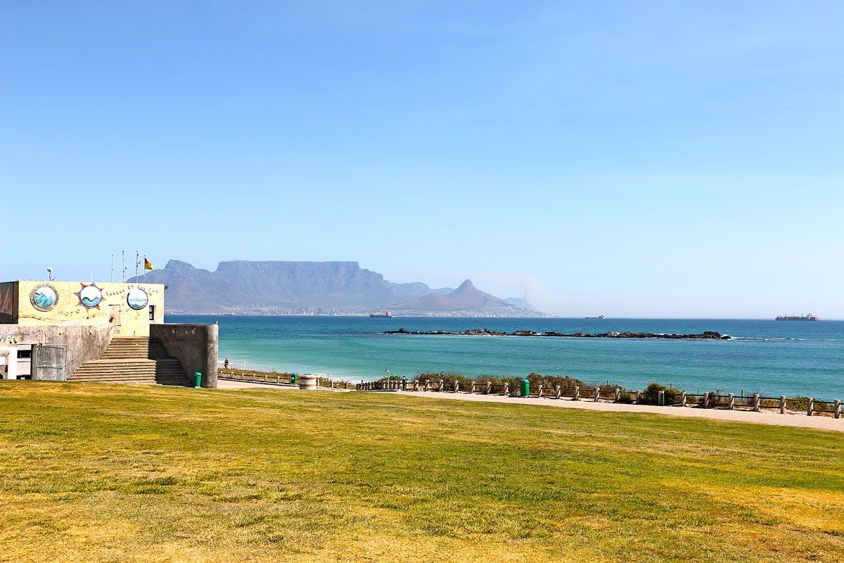 Vista costera con zona verde, un edificio con un mural y una cadena montañosa distante.