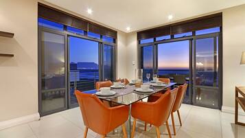 A dining area with a glass table and orange chairs, large windows with a city view, and a balcony with a railing.