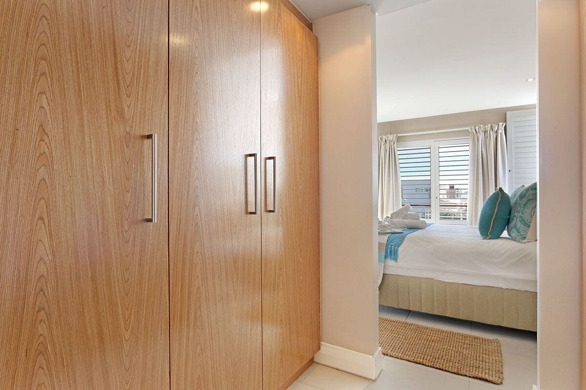 A bedroom with a large wooden wardrobe, a bed with a white duvet, and a view of the outside through a window with curtains.