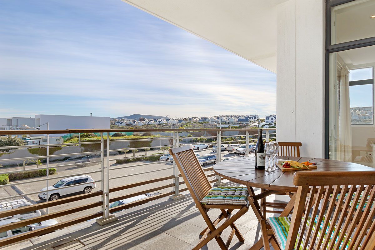 Un balcon avec une table et des chaises, donnant sur un parking et sur des bâtiments situés au loin.