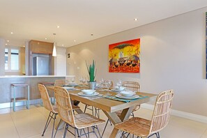 Uma sala de jantar com uma mesa de madeira posta para seis pessoas, rodeada por cadeiras de vime, uma cozinha ao fundo e um quadro abstrato vibrante na parede.