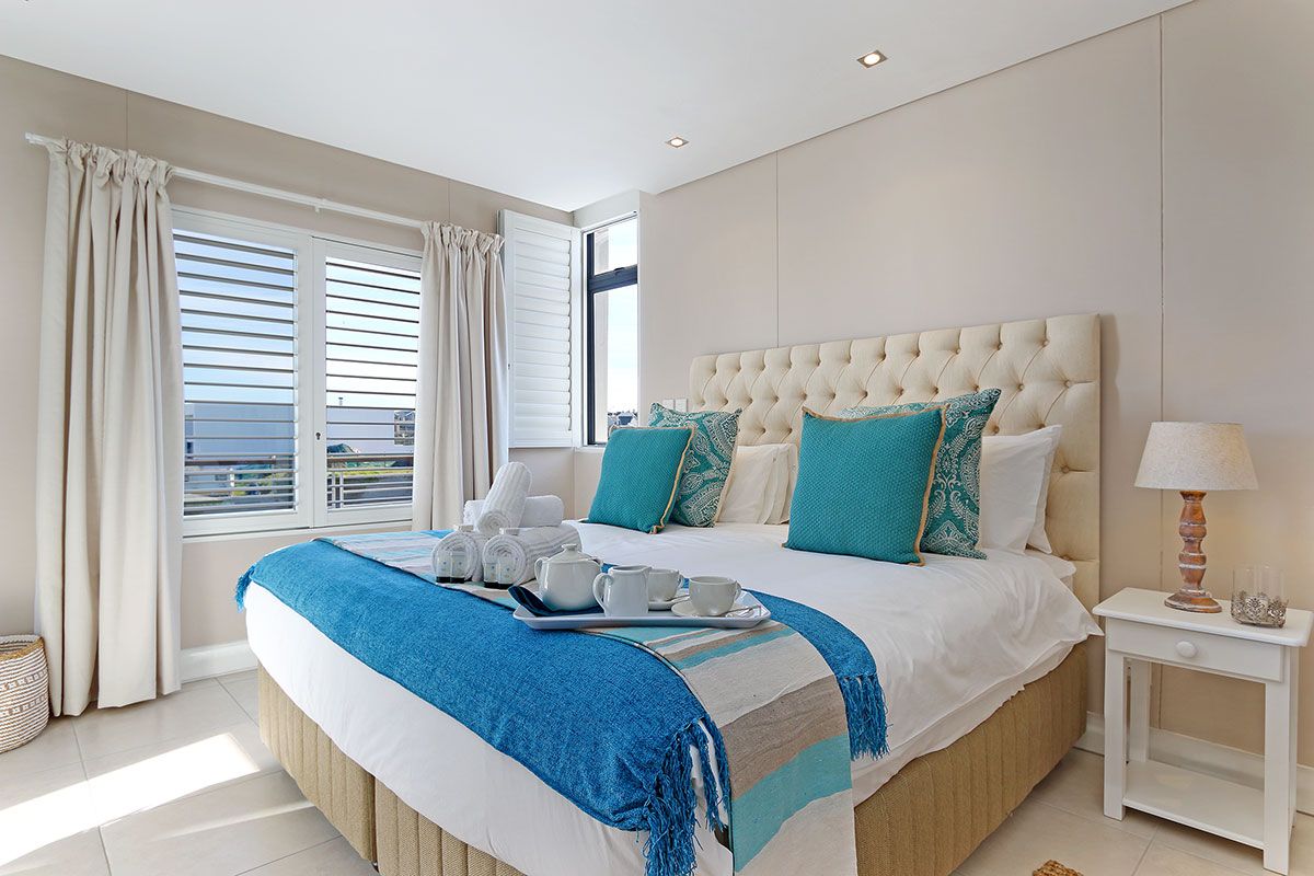 A bedroom with a large bed, a bedside table, a window with curtains, and a view of the sea.