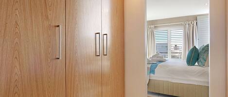 A bedroom with a large wooden wardrobe, a bed with a white duvet, and a view of the outside through a window with curtains.