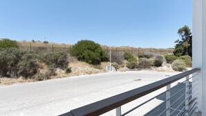 Une route asphaltée, avec une rambarde métallique au premier plan, mène à un paysage vallonné où la végétation est clairsemée et le ciel est d’un bleu limpide.