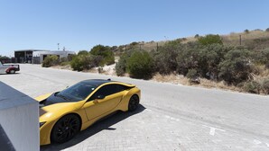 Une voiture de sport jaune est garée sur une route asphaltée, avec un bâtiment et une végétation desséchée en arrière-plan.