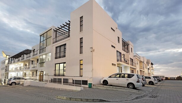 Un immeuble résidentiel à plusieurs étages doté de balcons, une aire de stationnement avec plusieurs voitures et un sol pavé.