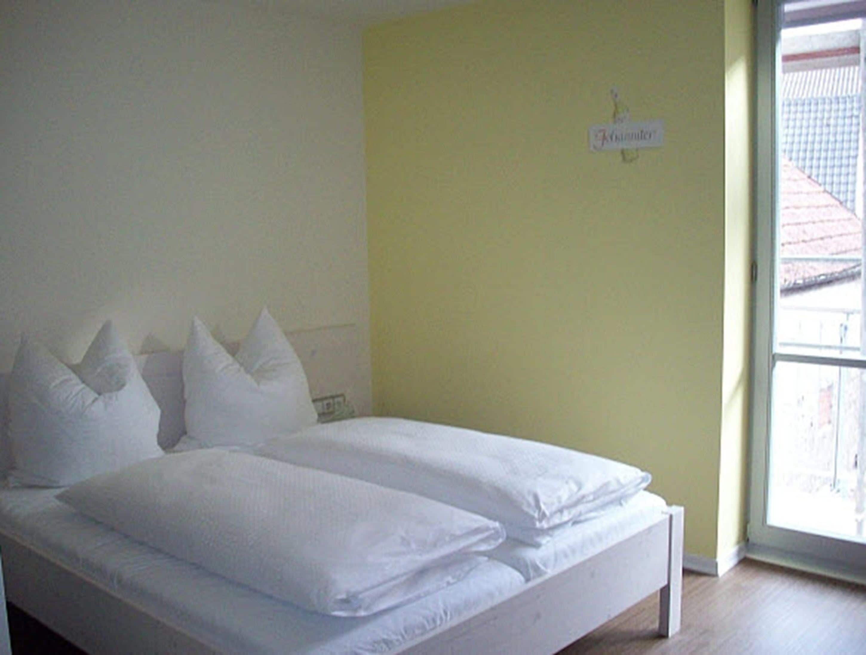 Un lit avec une literie et des oreillers blancs, un mur jaune et une fenêtre donnant sur un bâtiment à l’extérieur.