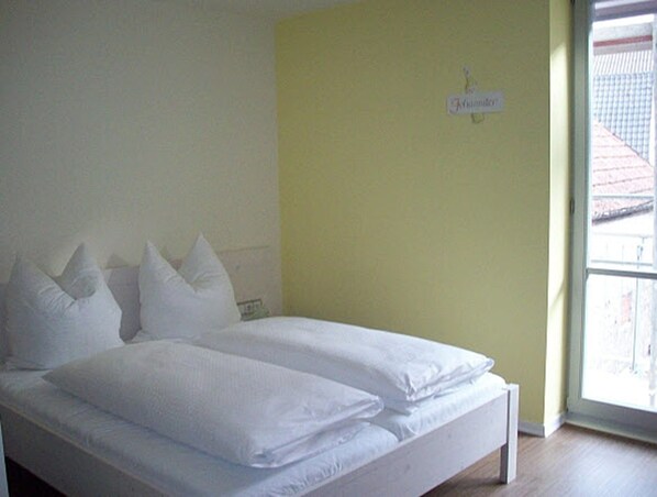 Una cama con ropa de cama y almohadas blancas, una pared amarilla y una ventana con vistas a un edificio.