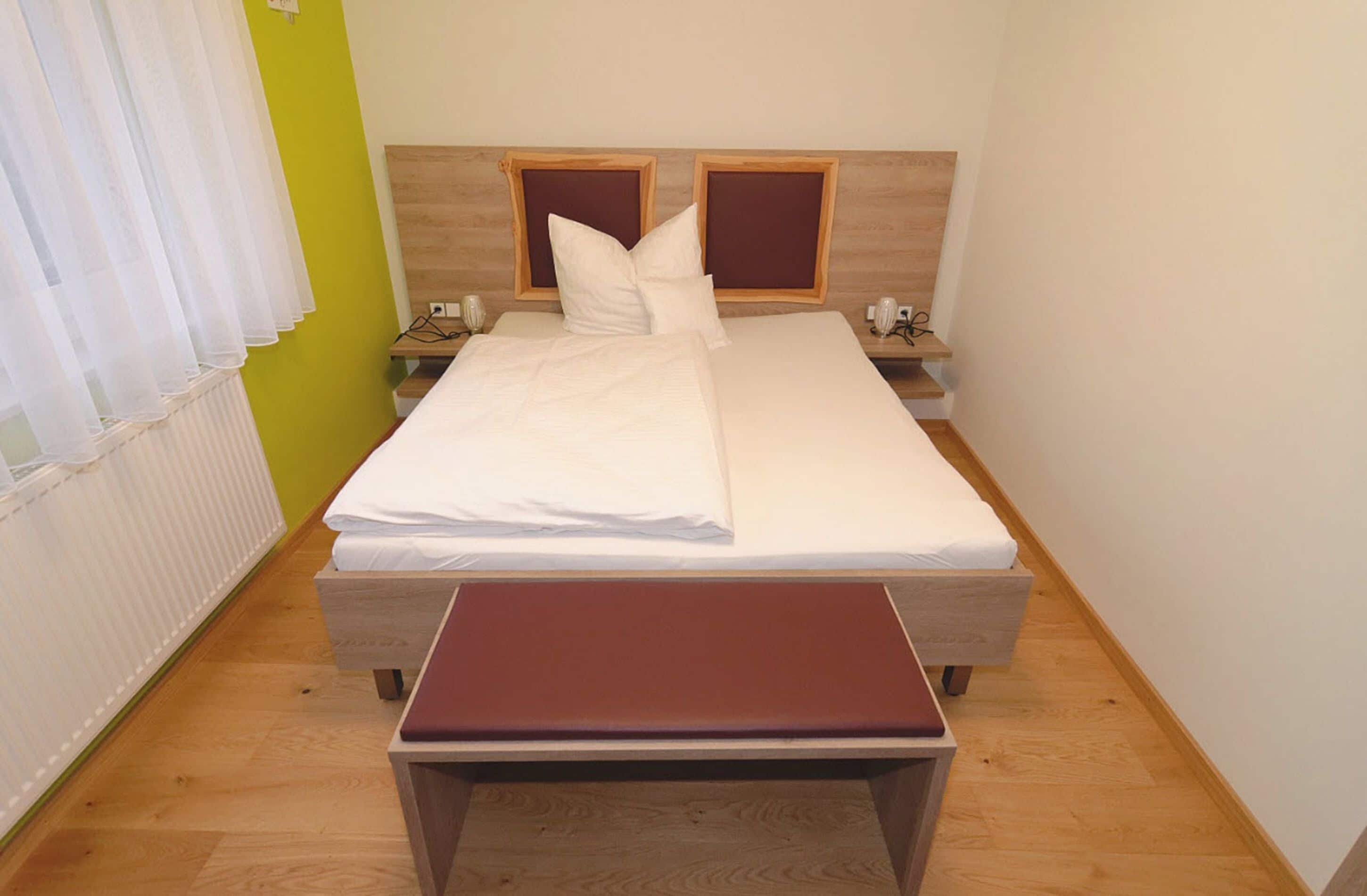 Ein ordentlich bezogenes Bett mit einem hölzernen Kopfteil, ein Nachttisch mit Lampe und eine Bank davor.
