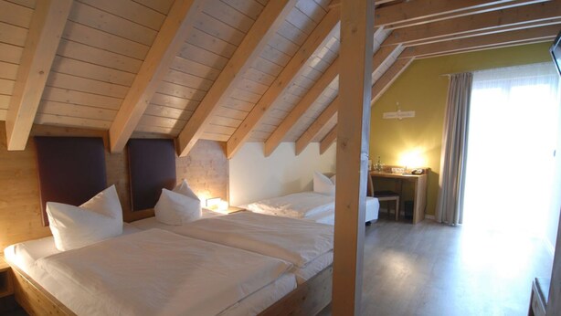 A bedroom with a wooden ceiling, a bed with white bedding, a nightstand, and a desk with a lamp. - RotHweinHotel (Wiesenbronn)