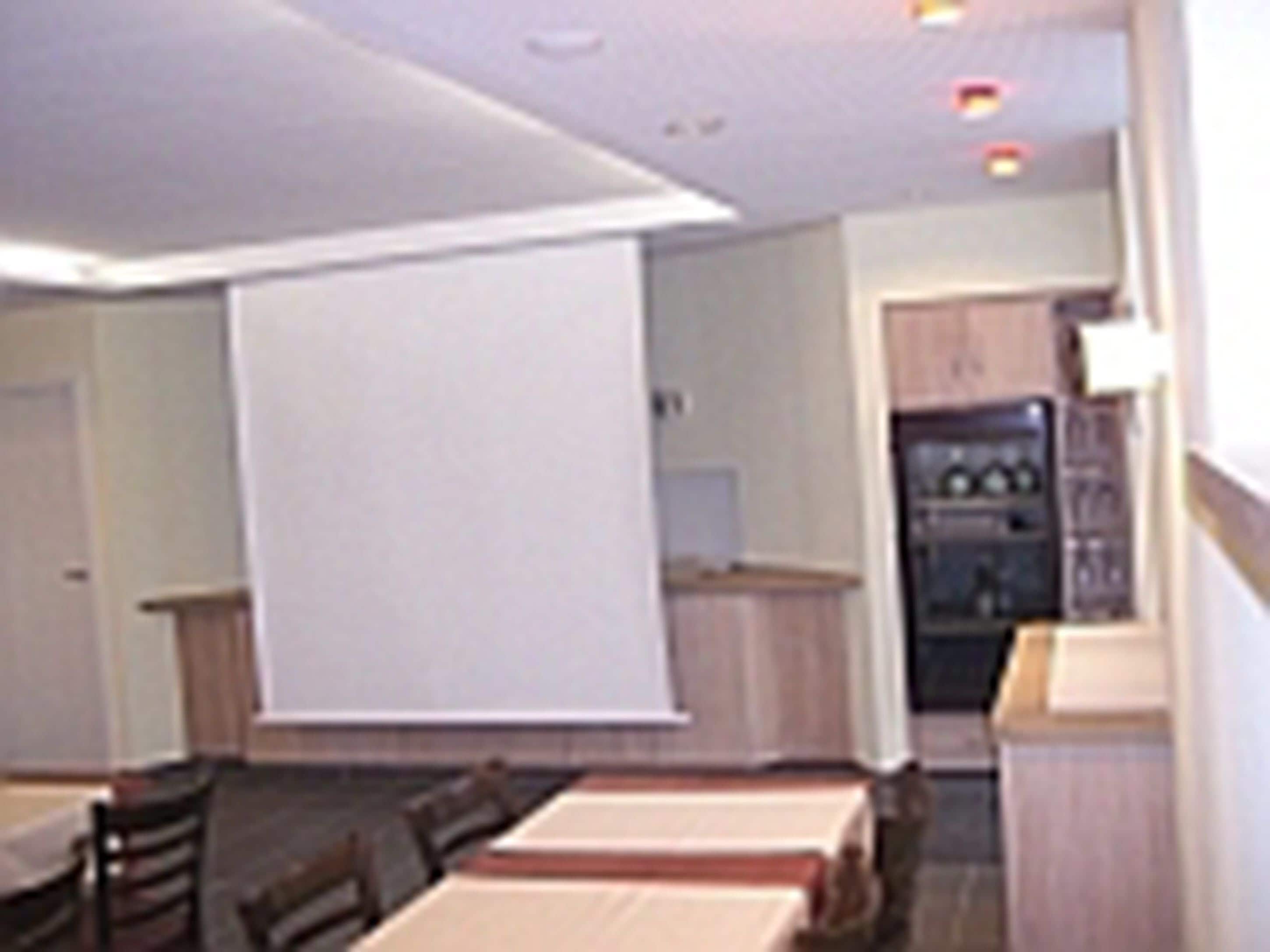 A room with a dining table, chairs, a projector screen, and a kitchenette in the background.