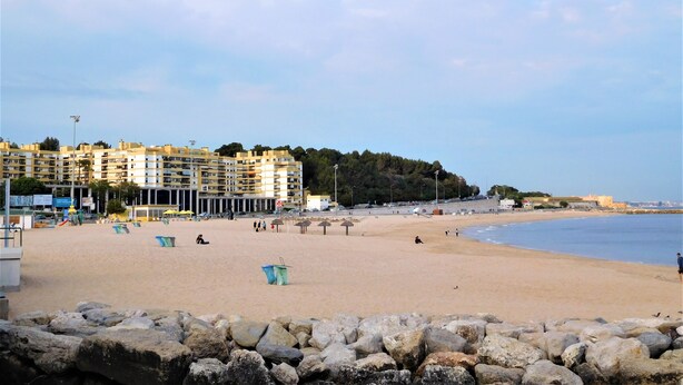 A beach with a row of multi-story buildings in the background, a sandy shore, and a rocky breakwater in the foreground. - Window to the Sea Oeiras (Oeiras)