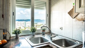 A kitchen with two sinks, a window with blinds, and a view of buildings and water. - Window to the Sea Oeiras (Oeiras)