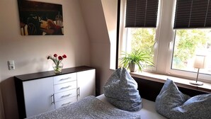 A bedroom with a bed, a dresser, a window with blinds, a plant, and a painting on the wall.
