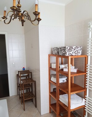 A bathroom with a wooden shelf holding towels, a chandelier, and a white radiator.