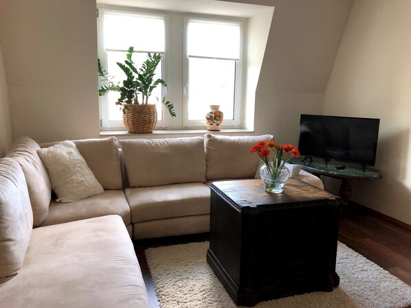 A living room with a beige sectional sofa, a small dark wooden coffee table with a vase of flowers, a television on a stand, and a window with two potted plants.