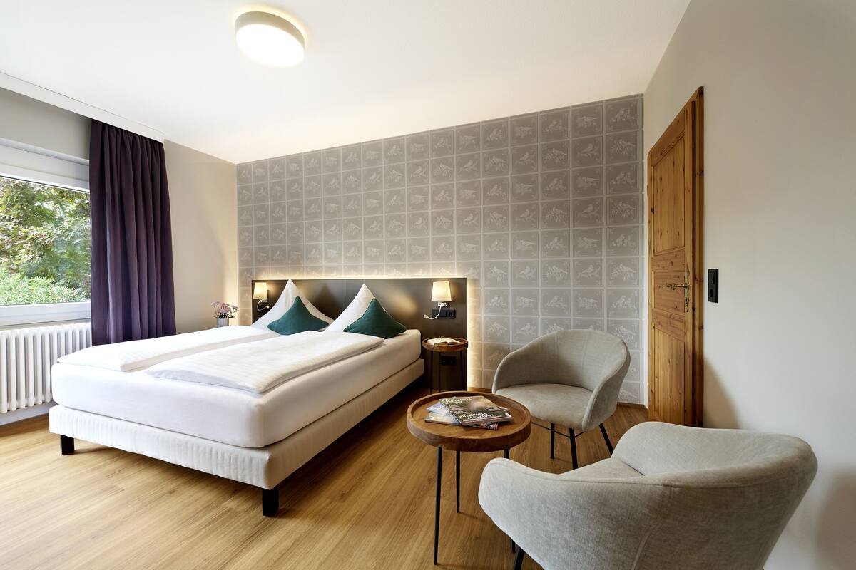 A hotel room with a bed, a round wooden table, a chair, and a nightstand with a lamp.
