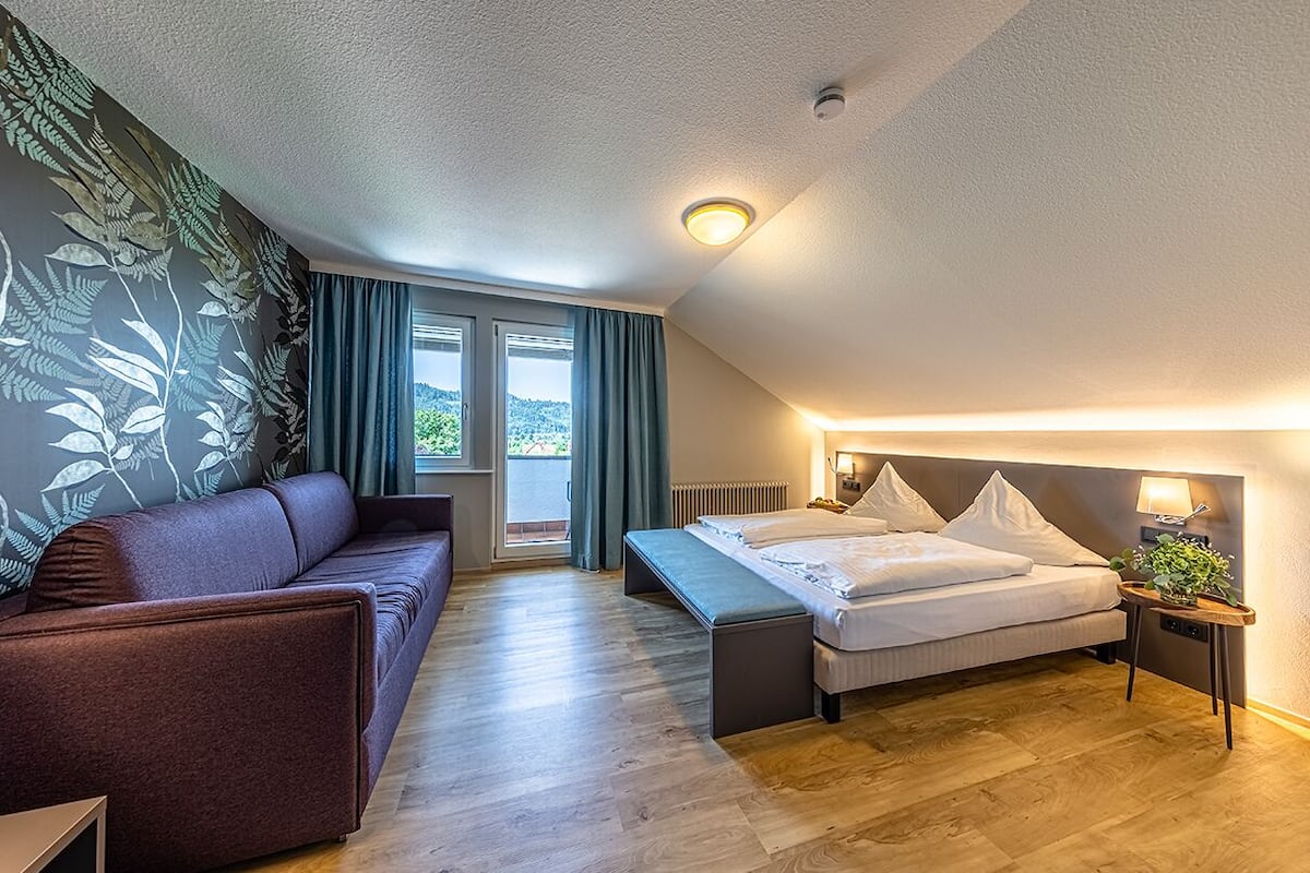 A modern hotel room with a bed, a sofa, a bench, and a view of the outdoors.