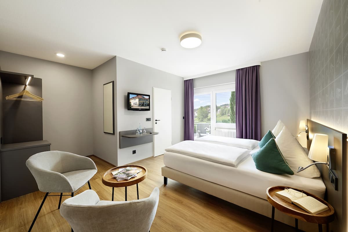 A modern hotel room with a sofa, two armchairs, a small table, a TV, a balcony with purple curtains, and a view of greenery.
